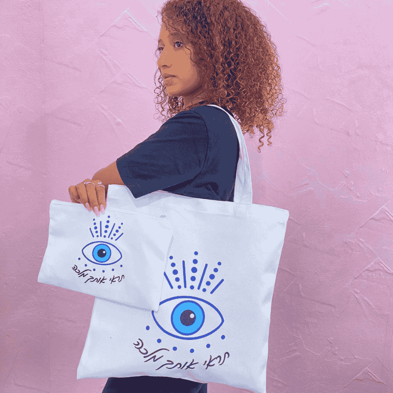 תיקי קנבס דגם תראי אותך מלכה כיתוב בצבע שחור תיק בצבע לבן ועליו ציור של עין כחולה
דוגמנית מדגמנת את התיקים
