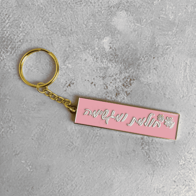 מחזיק מפתחות מוזהב דגם חולמת ומגשימה בכיתוב לבן עם ציור של פרח לבן על יד רקע ורוד